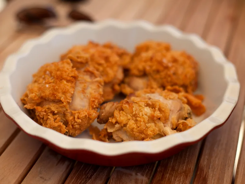 A fluted bowl of golden fried chicken pieces with a craggy, well-seasoned crust on a wooden table