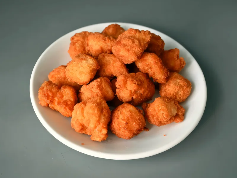 A white plate piled high with small, deep-golden breaded chicken nuggets with a rough craggy crust on a plain grey surface