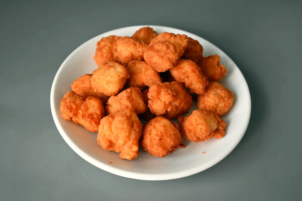 A white plate piled high with small, deep-golden breaded chicken nuggets with a rough craggy crust on a plain grey surface