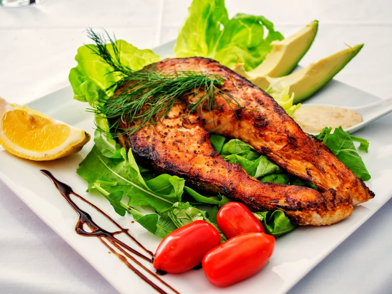 A crisp, deeply golden salmon steak on a bed of green leaves with lemon, cherry tomatoes, avocado and fresh dill