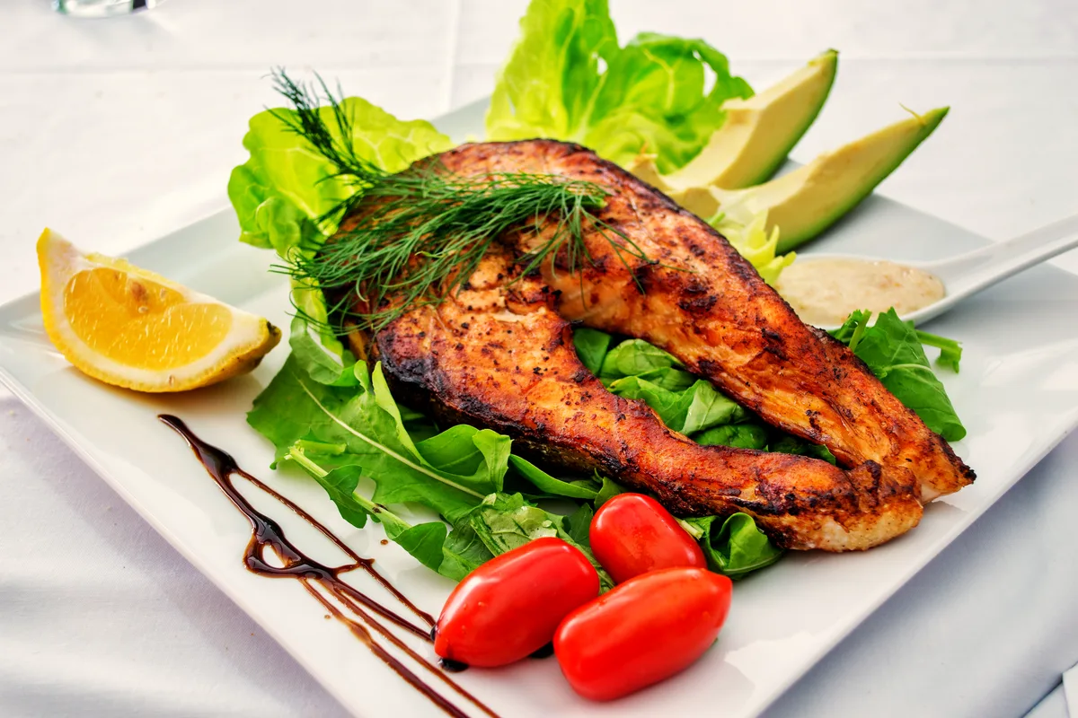 A crisp, deeply golden salmon steak on a bed of green leaves with lemon, cherry tomatoes, avocado and fresh dill