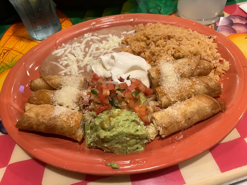Five golden crispy chicken taquitos dusted with grated cheese on a large terracotta plate with Mexican rice, refried beans with melted cheese, sour cream, pico de gallo and a scoop of guacamole in the centre