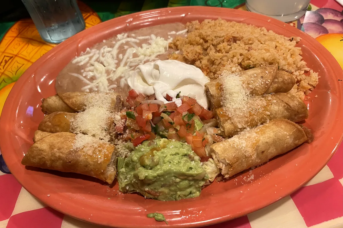 Five golden crispy chicken taquitos dusted with grated cheese on a large terracotta plate with Mexican rice, refried beans with melted cheese, sour cream, pico de gallo and a scoop of guacamole in the centre
