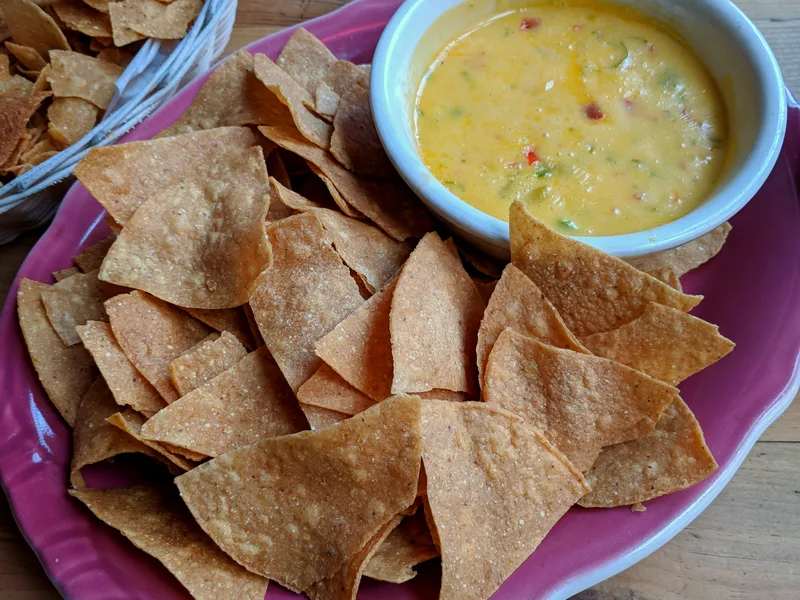 Golden air fryer tortilla chips on a pink plate with a bowl of warm queso dip and a basket of extra chips