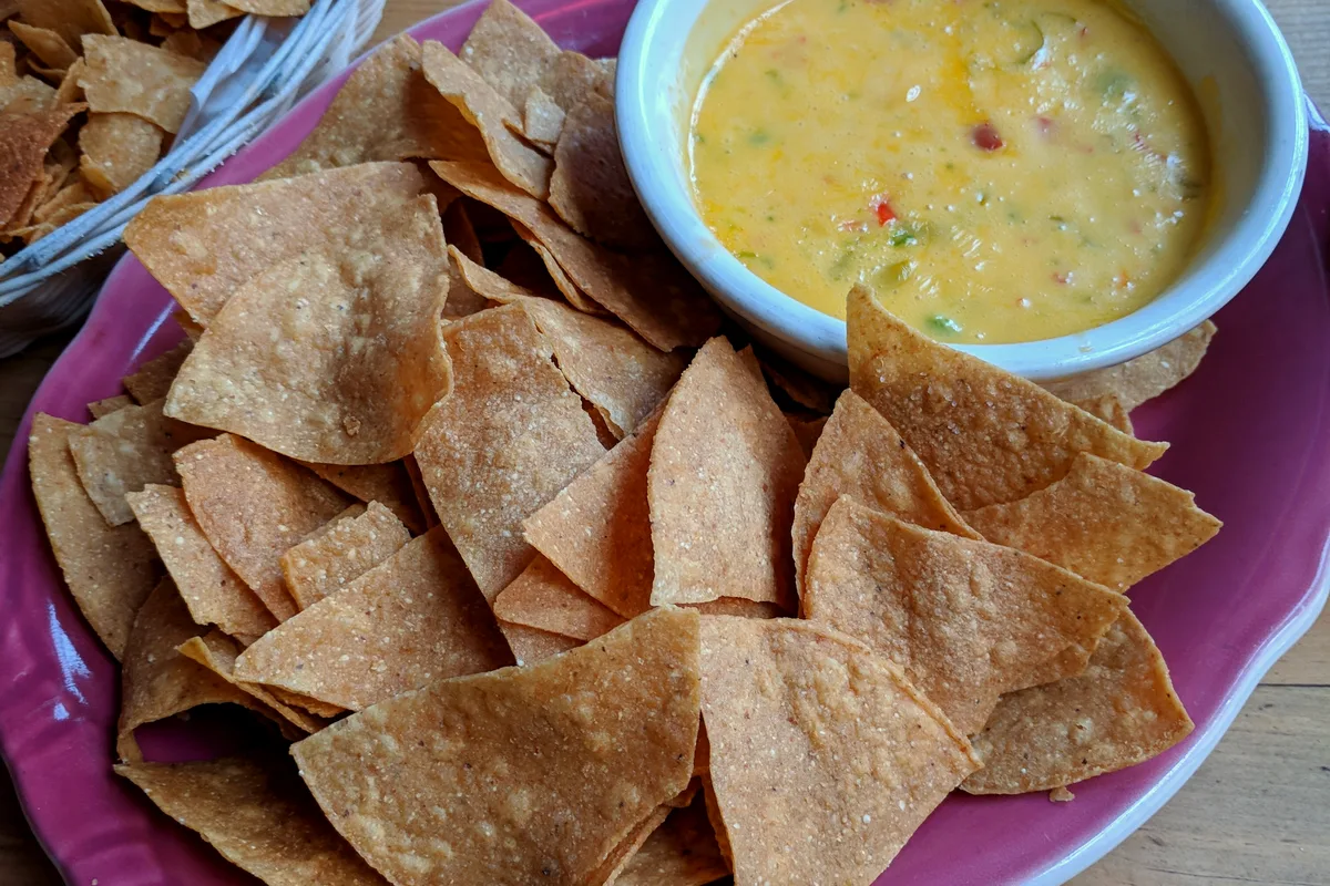 Golden air fryer tortilla chips on a pink plate with a bowl of warm queso dip and a basket of extra chips