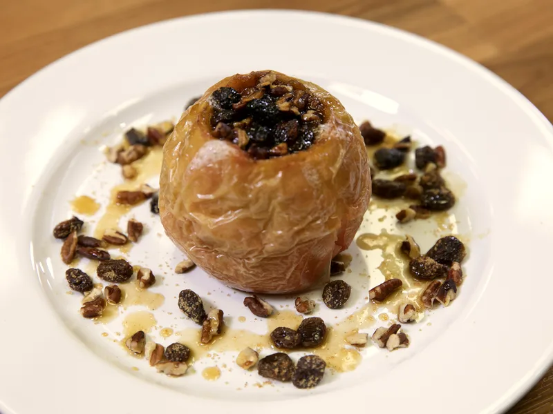A baked apple topped with chopped nuts and a warm syrup glaze on a white plate