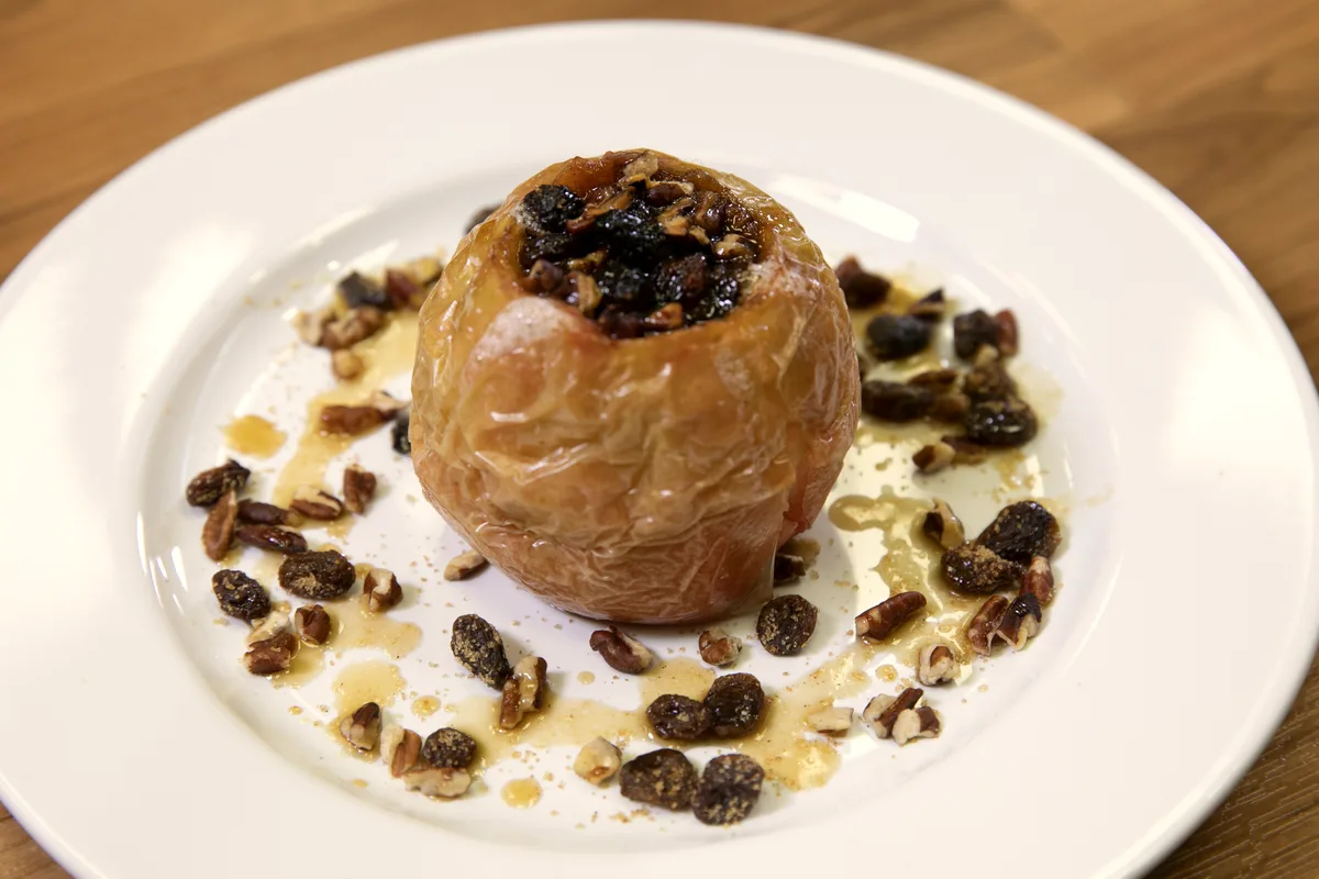 A baked apple topped with chopped nuts and a warm syrup glaze on a white plate