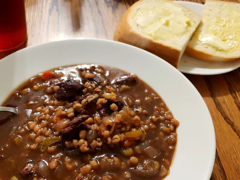 A white bowl of dark, rich beef and barley soup with tender beef chunks, plump pearl barley grains, onion and celery in a deep brown broth on a wooden table, with buttered white bread slices on a small plate behind and a glass of iced tea