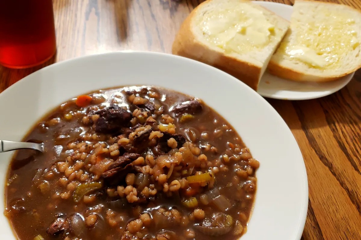 A white bowl of dark, rich beef and barley soup with tender beef chunks, plump pearl barley grains, onion and celery in a deep brown broth on a wooden table, with buttered white bread slices on a small plate behind and a glass of iced tea