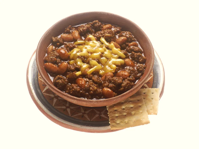 Bowl of beef chili with beans topped with melted cheese, served with crackers