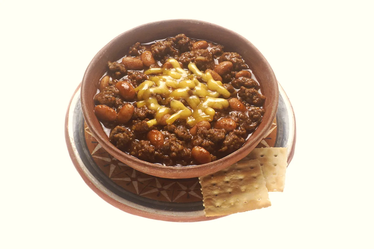 Bowl of beef chili with beans topped with melted cheese, served with crackers