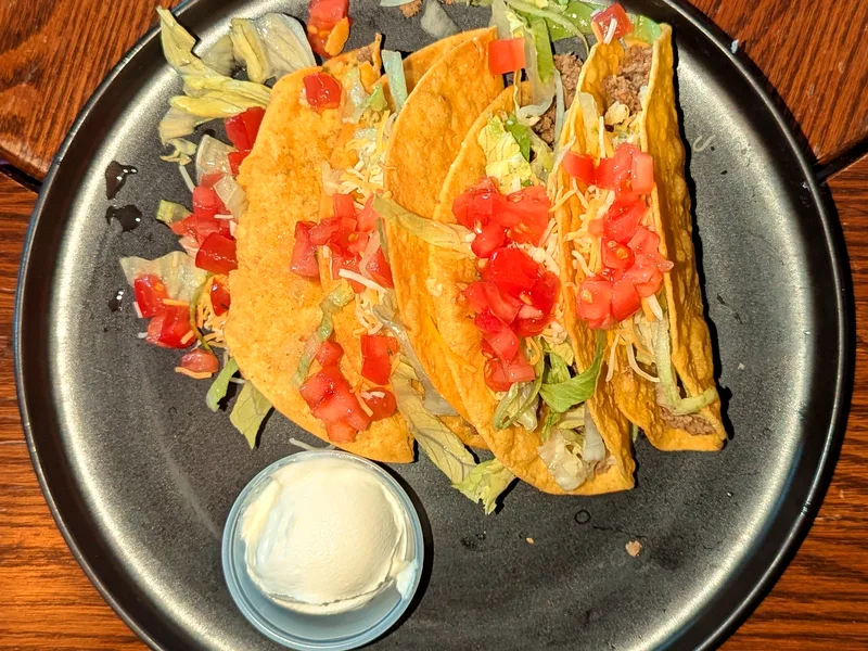 Four crunchy corn tacos filled with seasoned ground beef, shredded lettuce, diced tomato and cheese on a dark plate, with a small pot of sour cream