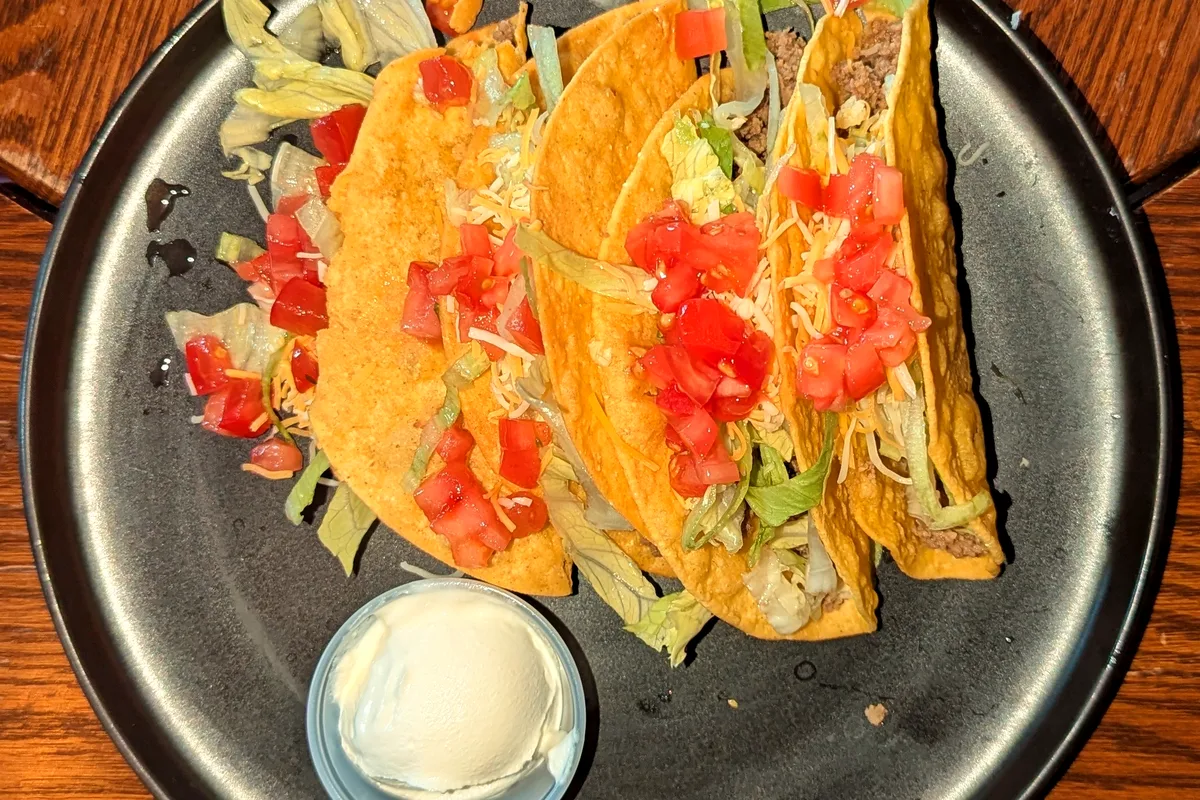 Four crunchy corn tacos filled with seasoned ground beef, shredded lettuce, diced tomato and cheese on a dark plate, with a small pot of sour cream