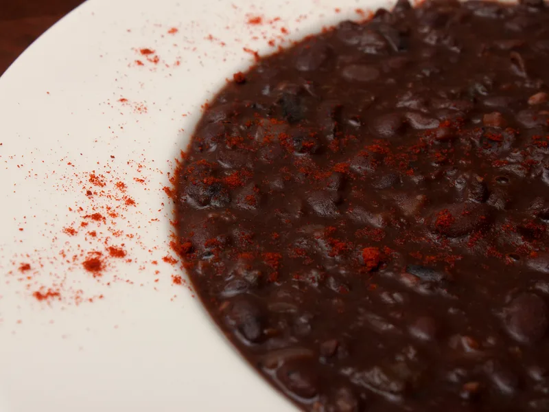 A close-up of thick black bean soup in a white bowl, whole beans visible in the dark, glossy broth with a dusting of red paprika over the top