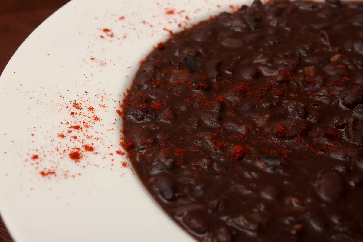 A close-up of thick black bean soup in a white bowl, whole beans visible in the dark, glossy broth with a dusting of red paprika over the top