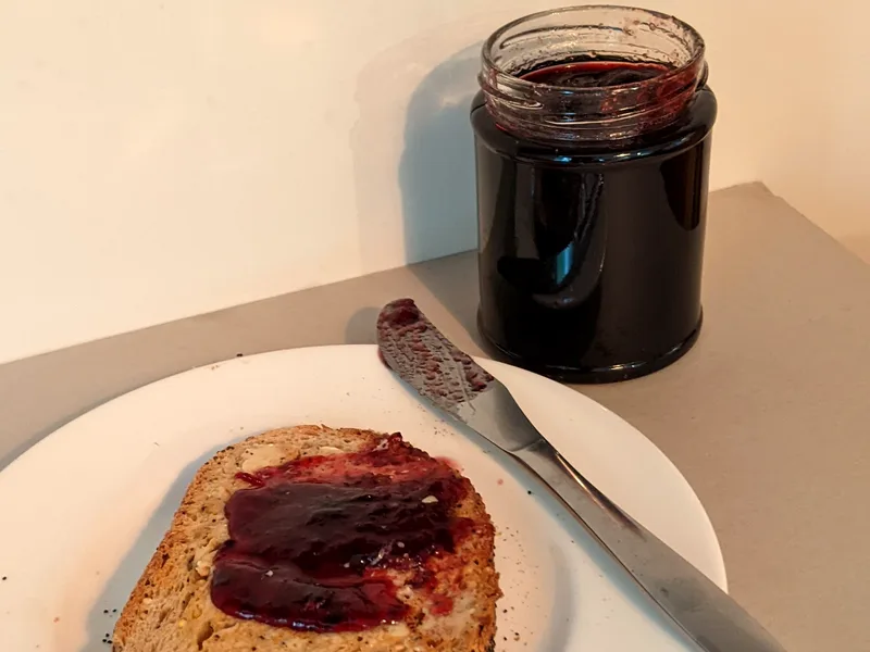 An open jar of deep purple blackberry jam beside a slice of seeded toast spread thickly with the jam on a white plate with a butter knife