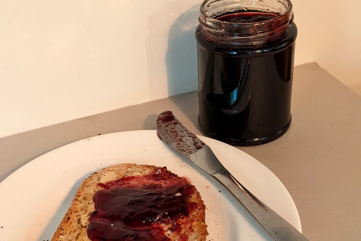 An open jar of deep purple blackberry jam beside a slice of seeded toast spread thickly with the jam on a white plate with a butter knife
