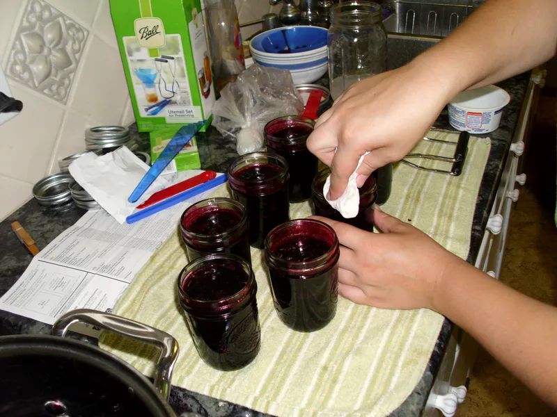 Open jars of deep purple blueberry jam being wiped clean before lids go on for canning