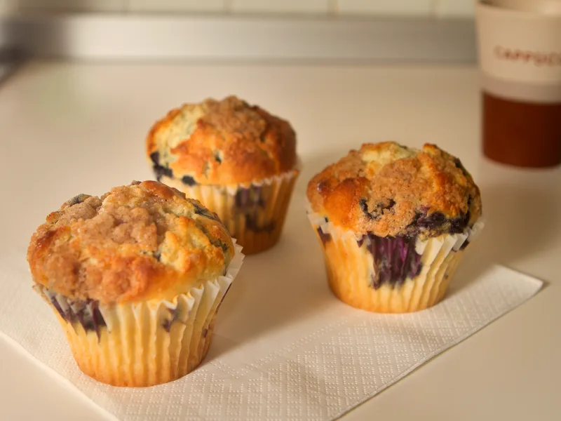 Three blueberry muffins in paper liners with golden streusel tops and berries bursting through, a cup of coffee behind