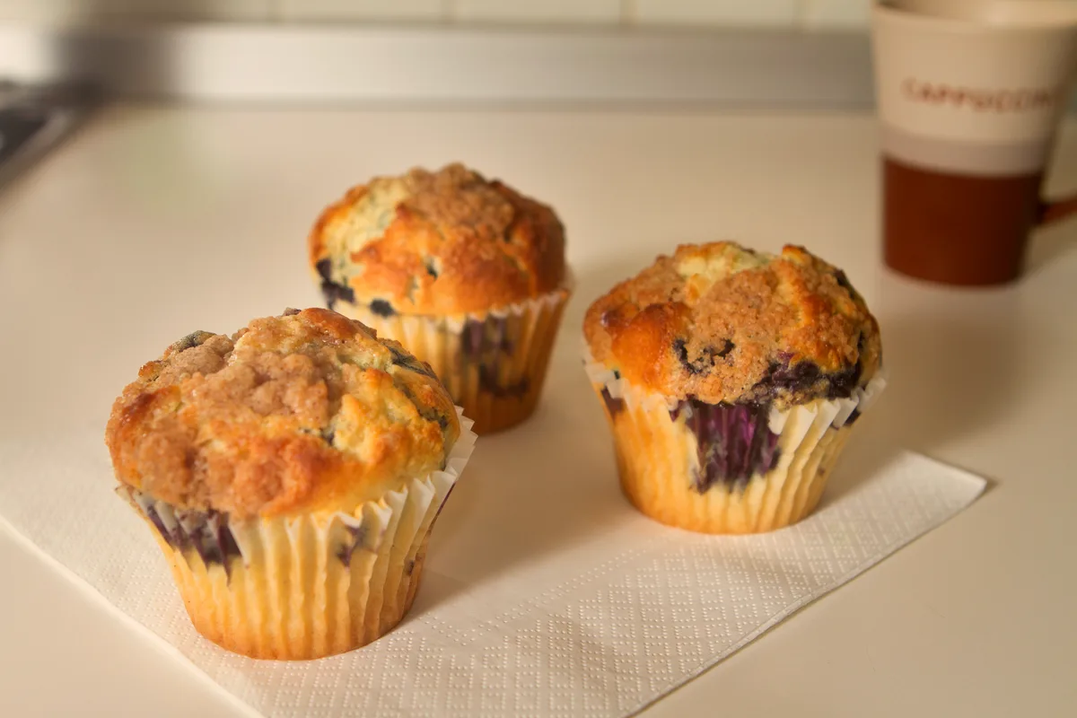 Three blueberry muffins in paper liners with golden streusel tops and berries bursting through, a cup of coffee behind