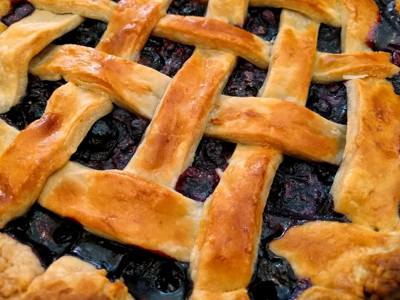 A close-up of blueberry pie with a golden woven lattice crust and dark, jammy blueberries bubbling between the strips
