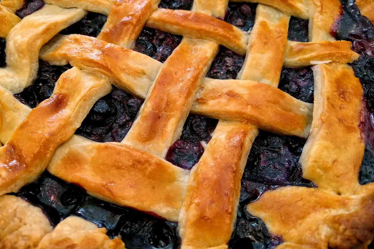 A close-up of blueberry pie with a golden woven lattice crust and dark, jammy blueberries bubbling between the strips
