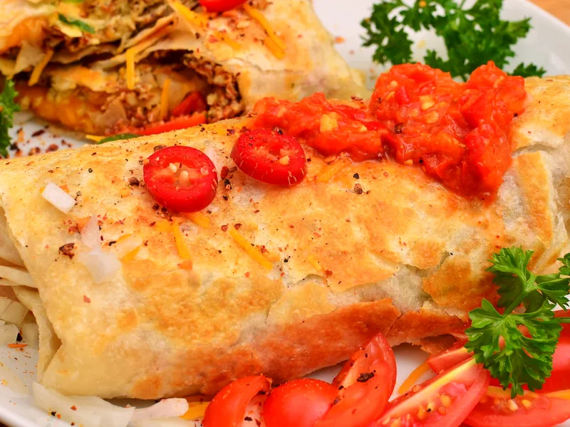 Toasted breakfast burrito topped with red pepper salsa and sliced chiles, a halved burrito behind it showing the egg and cheese filling, cherry tomatoes alongside