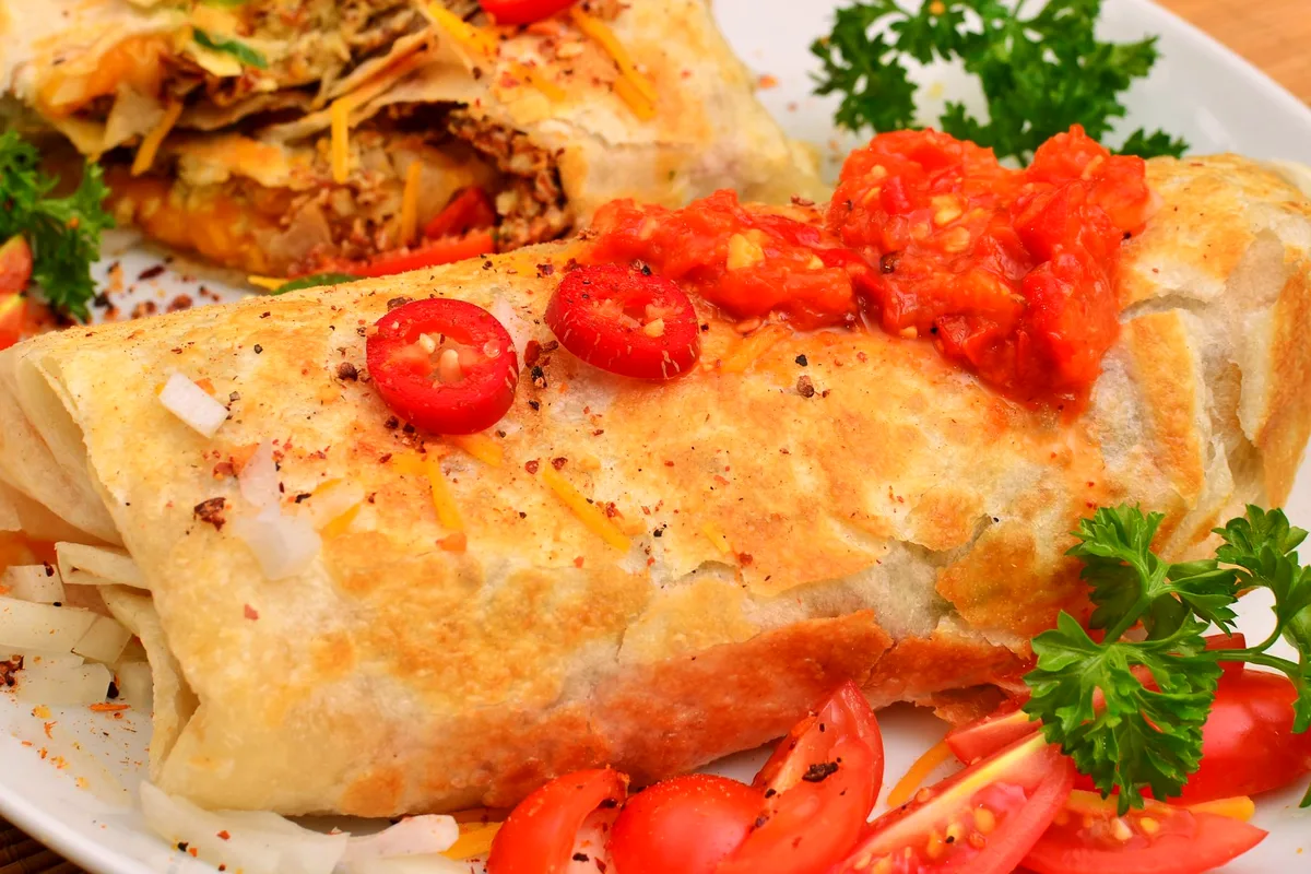 Toasted breakfast burrito topped with red pepper salsa and sliced chiles, a halved burrito behind it showing the egg and cheese filling, cherry tomatoes alongside