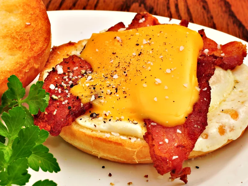 An open bacon, egg and cheese breakfast sandwich on a toasted roll, with a draped slice of melted cheddar over crisp bacon and a fried egg, dusted with flaky salt and black pepper, the toasted top leaning alongside and a parsley sprig in front