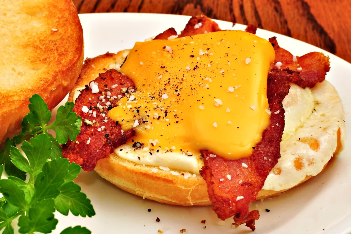 An open bacon, egg and cheese breakfast sandwich on a toasted roll, with a draped slice of melted cheddar over crisp bacon and a fried egg, dusted with flaky salt and black pepper, the toasted top leaning alongside and a parsley sprig in front