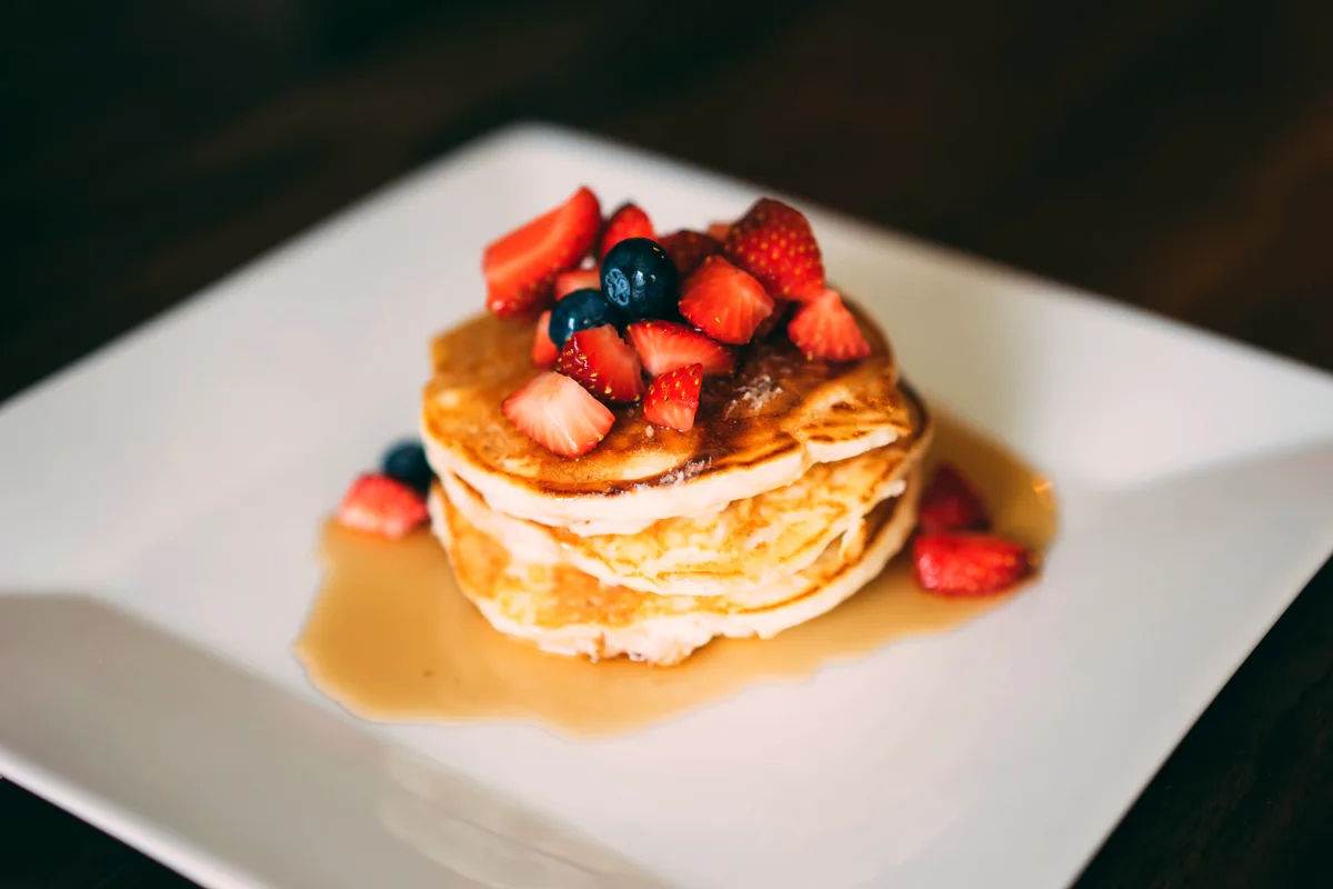 Stack of fluffy buttermilk pancakes topped with strawberries and blueberries with syrup on a white plate