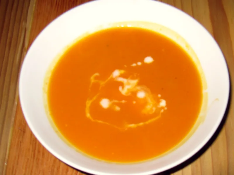 A white bowl of smooth golden-orange butternut squash soup with a swirl of cream on top, seen from above on a wooden table