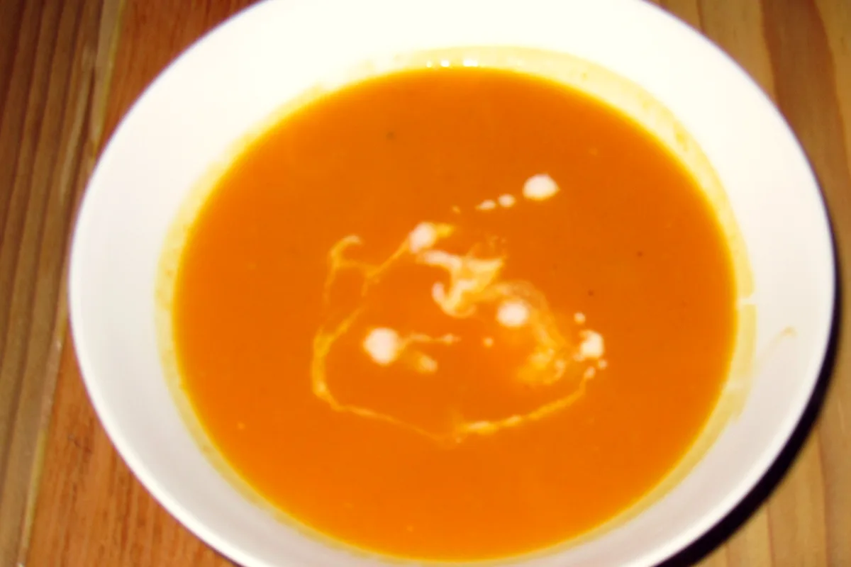 A white bowl of smooth golden-orange butternut squash soup with a swirl of cream on top, seen from above on a wooden table
