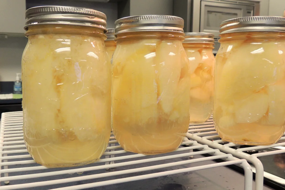 Sealed glass jars of pale golden canned pear pieces in light syrup cooling on a white wire rack