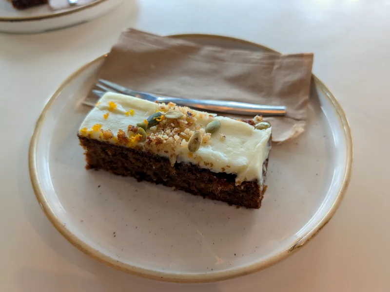 A slice of spiced carrot cake topped with white cream cheese frosting, chopped nuts and seeds on a speckled plate