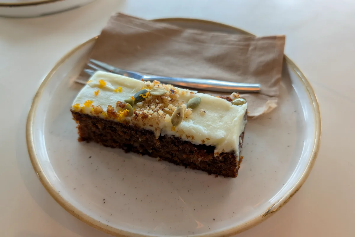 A slice of spiced carrot cake topped with white cream cheese frosting, chopped nuts and seeds on a speckled plate