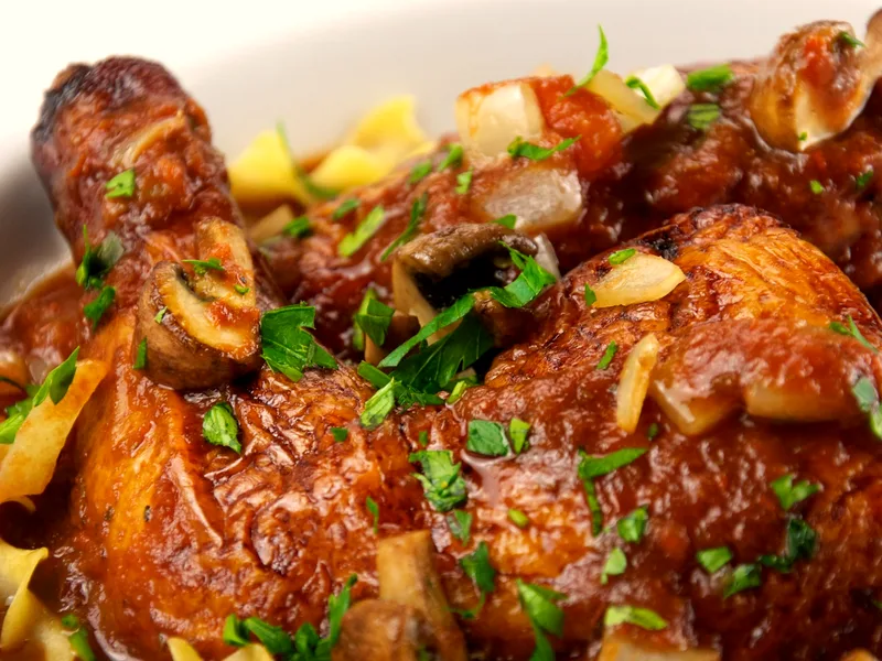 Chicken cacciatore with braised chicken in rich tomato sauce with mushrooms, diced onion and parsley, served over egg noodles in a white bowl