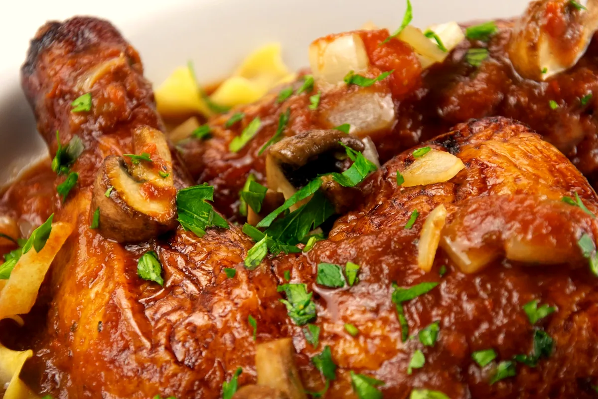 Chicken cacciatore with braised chicken in rich tomato sauce with mushrooms, diced onion and parsley, served over egg noodles in a white bowl