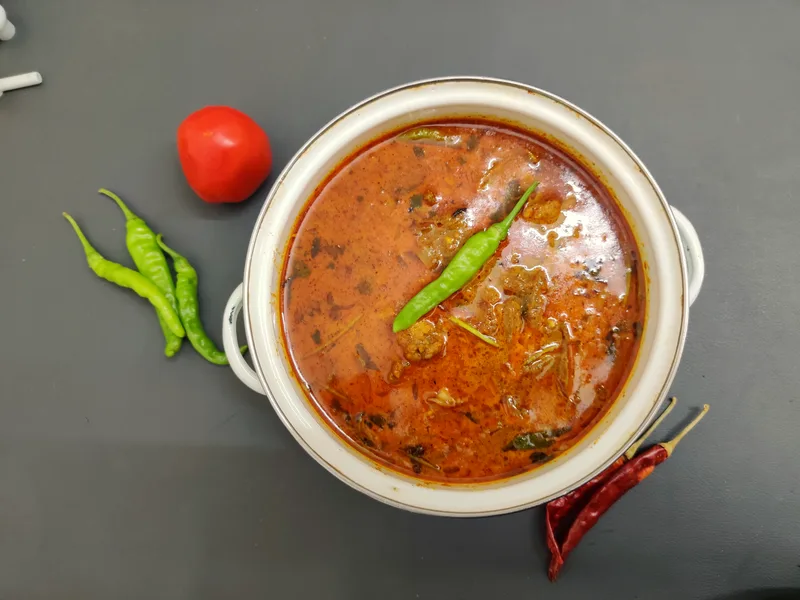 Slow cooker chicken curry in a white pot, chicken in a rich tomato-coconut sauce topped with a green chili, with a tomato and dried chilies beside it