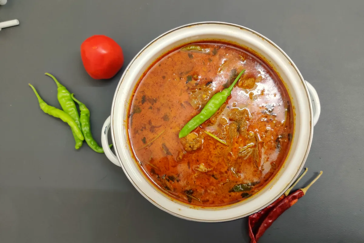 Slow cooker chicken curry in a white pot, chicken in a rich tomato-coconut sauce topped with a green chili, with a tomato and dried chilies beside it