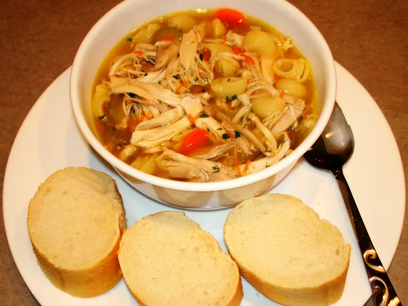 Bowl of chicken noodle soup with shredded chicken, shell pasta and carrots, sliced bread alongside