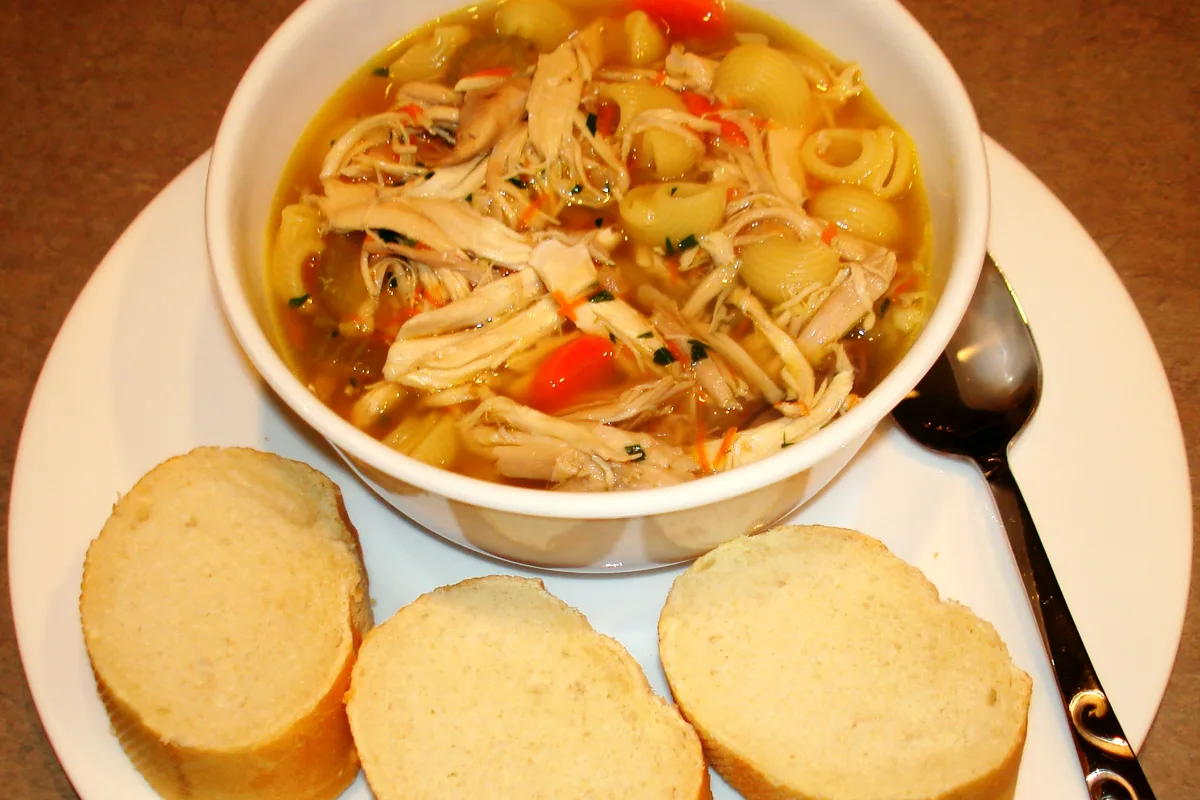 Bowl of chicken noodle soup with shredded chicken, shell pasta and carrots, sliced bread alongside