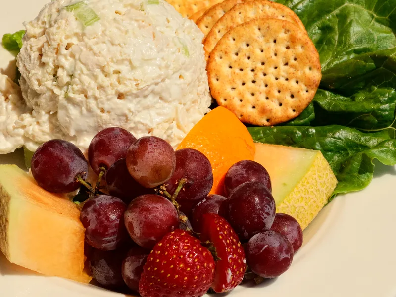 Scoop of creamy chicken salad with celery on lettuce, served with crackers and fresh fruit alongside