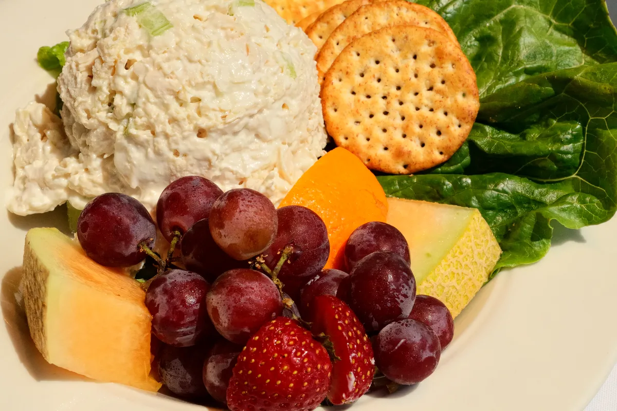 Scoop of creamy chicken salad with celery on lettuce, served with crackers and fresh fruit alongside