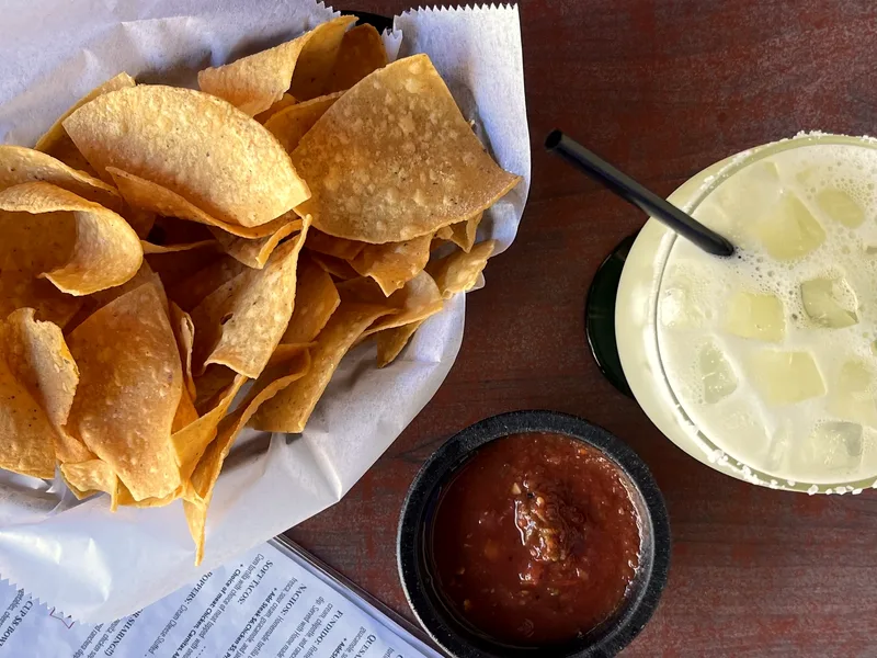 A basket of golden tortilla chips on white paper next to a small dark bowl of red tomato salsa on a wooden table
