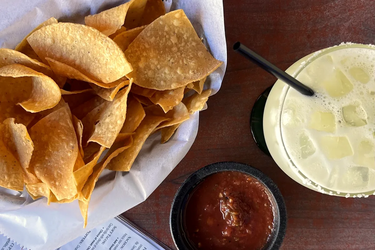 A basket of golden tortilla chips on white paper next to a small dark bowl of red tomato salsa on a wooden table