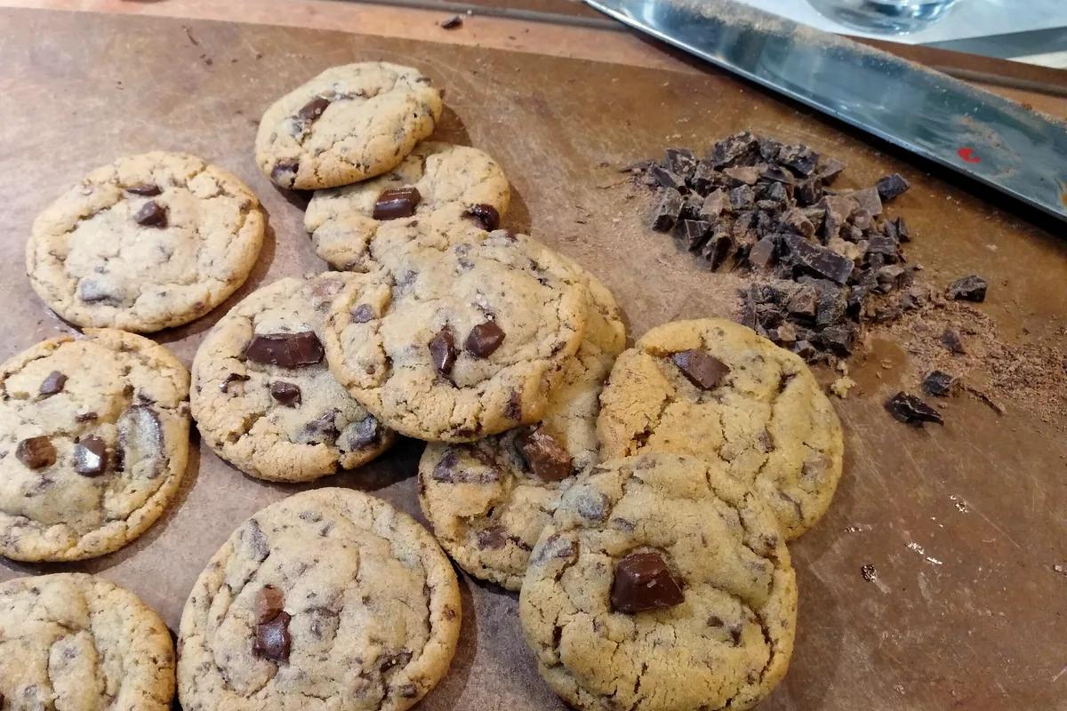 Freshly baked chocolate chip cookies with chocolate chunks cooling on a wooden board next to chopped chocolate