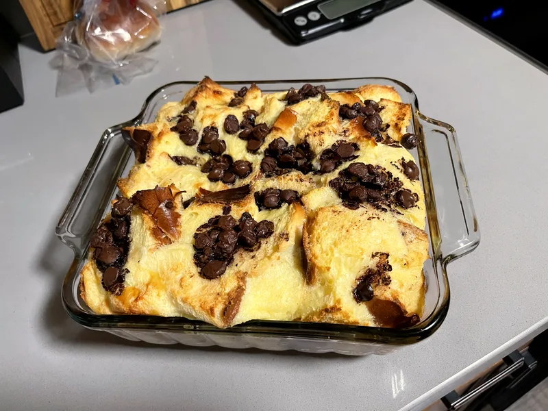 Chocolate chip bread pudding baked golden in a clear glass baking dish, custardy brioche cubes studded with melted chocolate chips