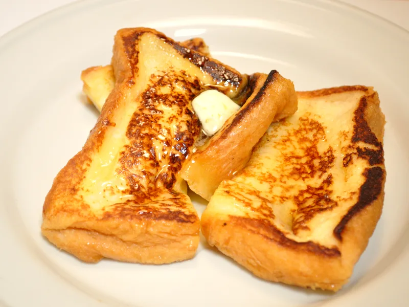 Thick slices of golden French toast with browned custard spots, melting butter and syrup on a white plate