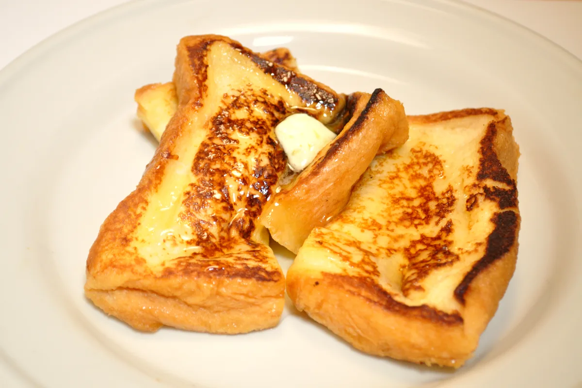 Thick slices of golden French toast with browned custard spots, melting butter and syrup on a white plate